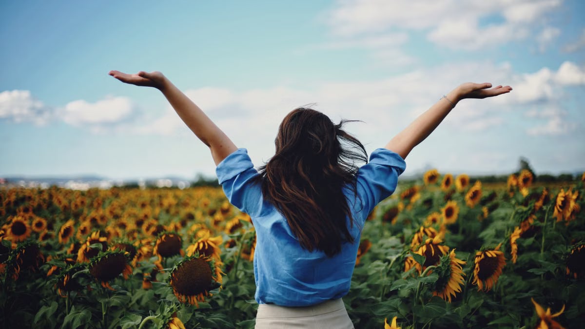 A woman in a blue shirt joyfully raises her arms amidst a sunflower field in Izmir, Türkiye.