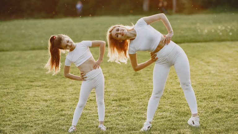 A mother and daughter in activewear stretching on a sunny day in a grassy field.