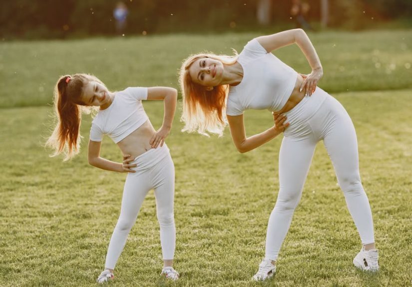 A mother and daughter in activewear stretching on a sunny day in a grassy field.