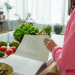 A woman in a kitchen flipping through a cookbook surrounded by fresh vegetables.