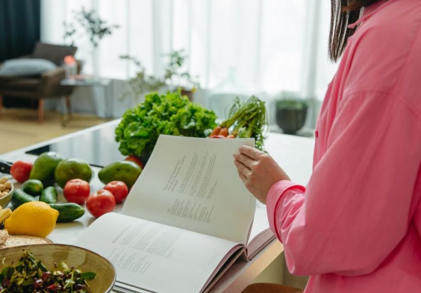 A woman in a kitchen flipping through a cookbook surrounded by fresh vegetables.