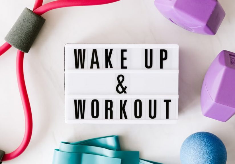 Top view closeup composition of wake up and workout inscription on light box surface among expanders and dumbbells and massage ball on white background